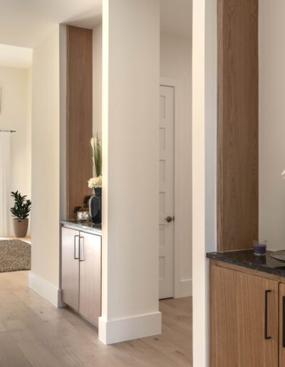 Modern hallway with light wood floors, built-in cabinets, a dark vase with flowers, and a view into a bright living area with large windows and a small table.