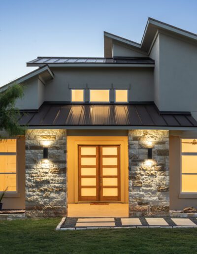 Modern house exterior with illuminated stone facade, double wooden front door, large windows, and geometric pathway at dusk.